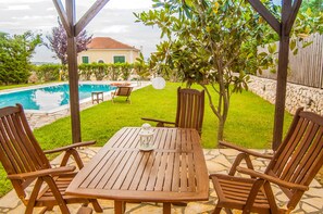 Eine Terrasse im Freien mit einem Holztisch und -stühlen, einer Laterne und Blick auf Pool und Haus im Hintergrund.