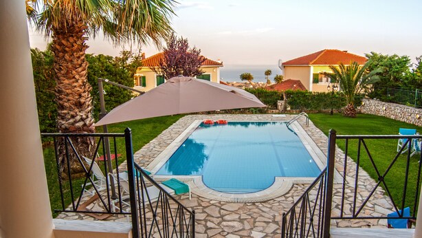 A swimming pool with a stone deck, surrounded by a metal railing, and a large umbrella providing shade.