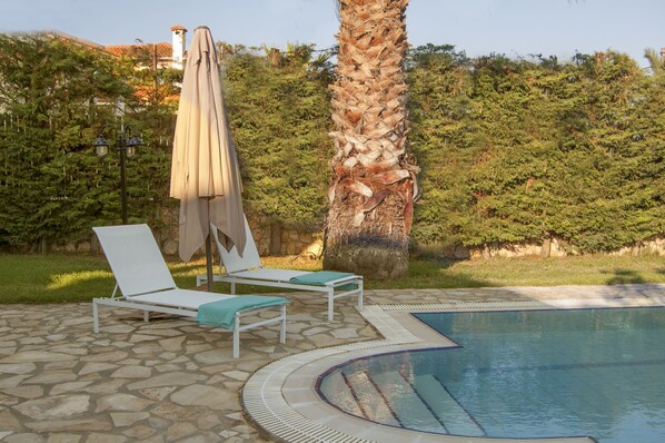 Un espace piscine agrémenté de chaises longues et d’un parasol, entouré de verdure et d’un mur en pierre.