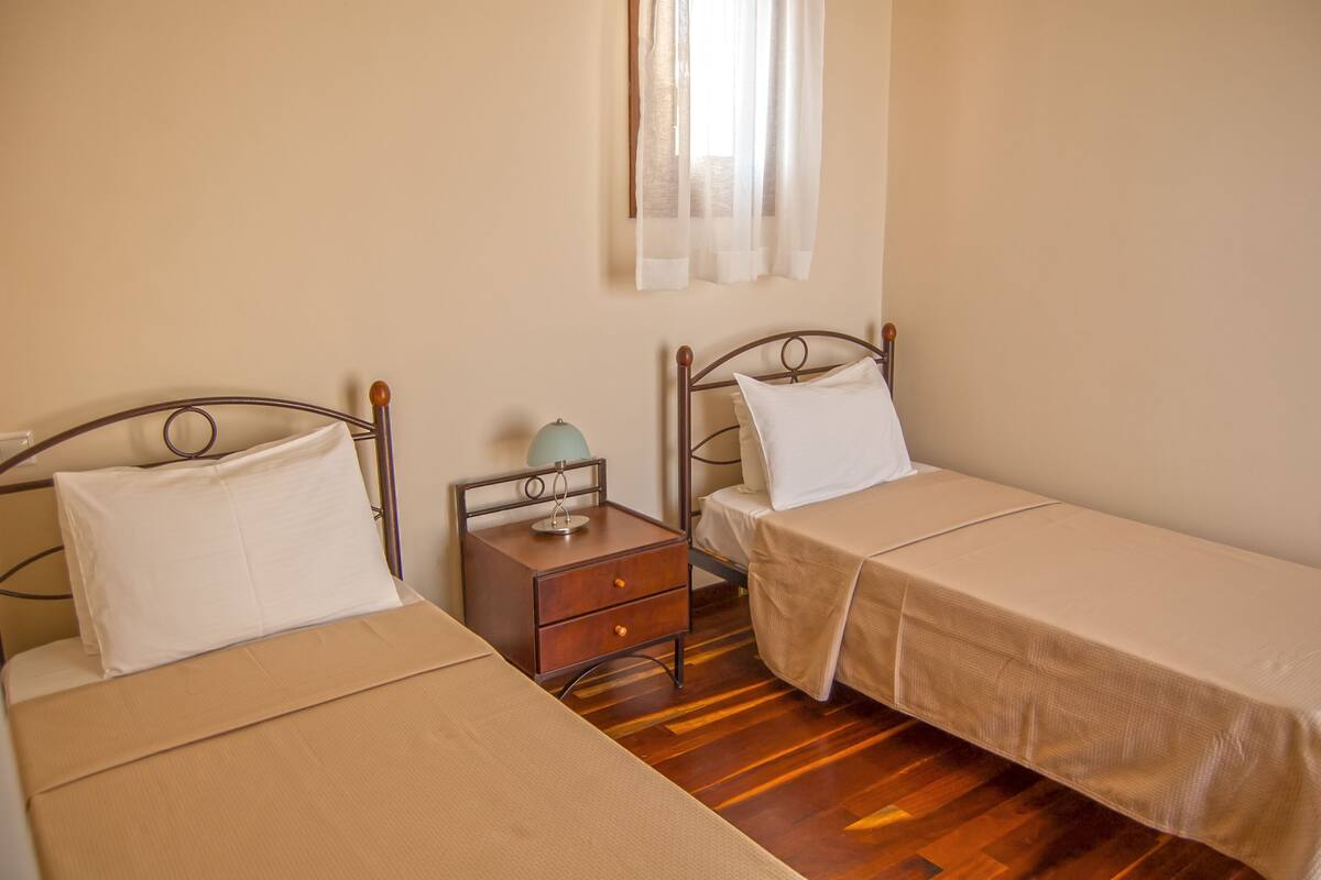 A bedroom with two single beds, a nightstand with a lamp, and a window with sheer curtains.