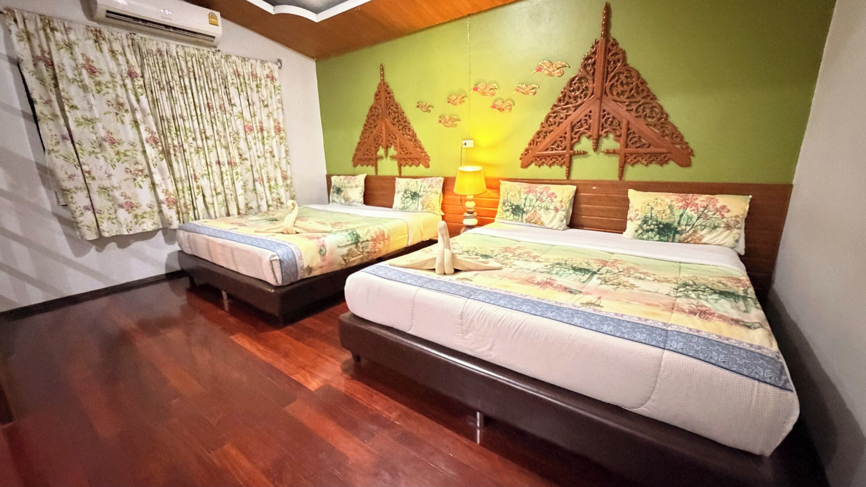 A hotel room with two beds, a wooden floor, a green wall with decorative elements, and an air conditioning unit.