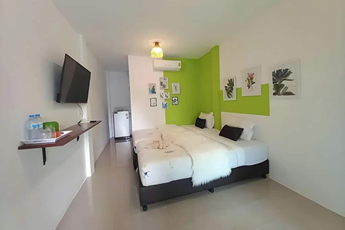 Un dormitorio con una cama, un televisor montado en la pared, un refrigerador pequeño y una estantería con objetos.