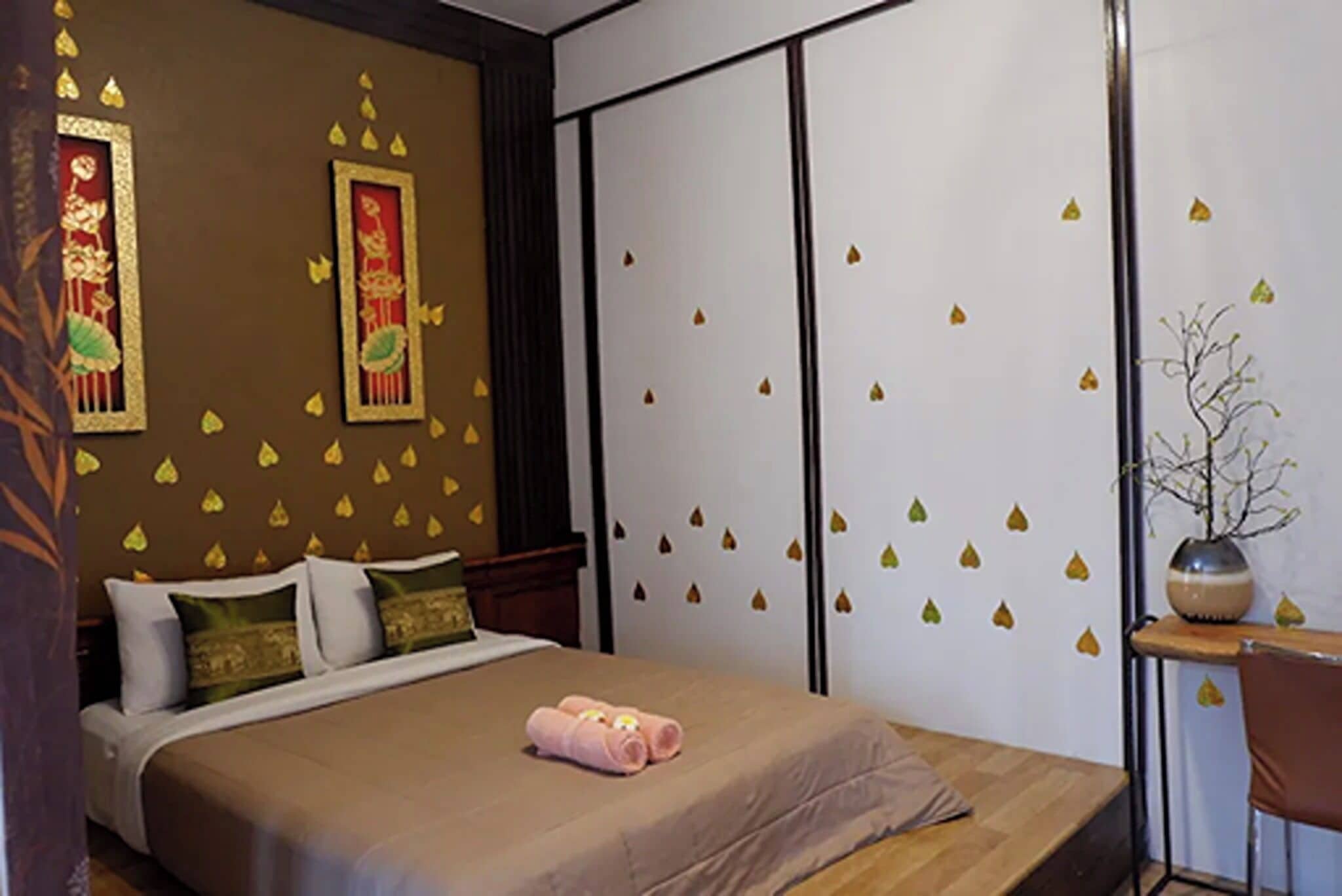 A hotel room with a bed, pillows, a bedside table with a vase, and a wall with decorative leaf decals.