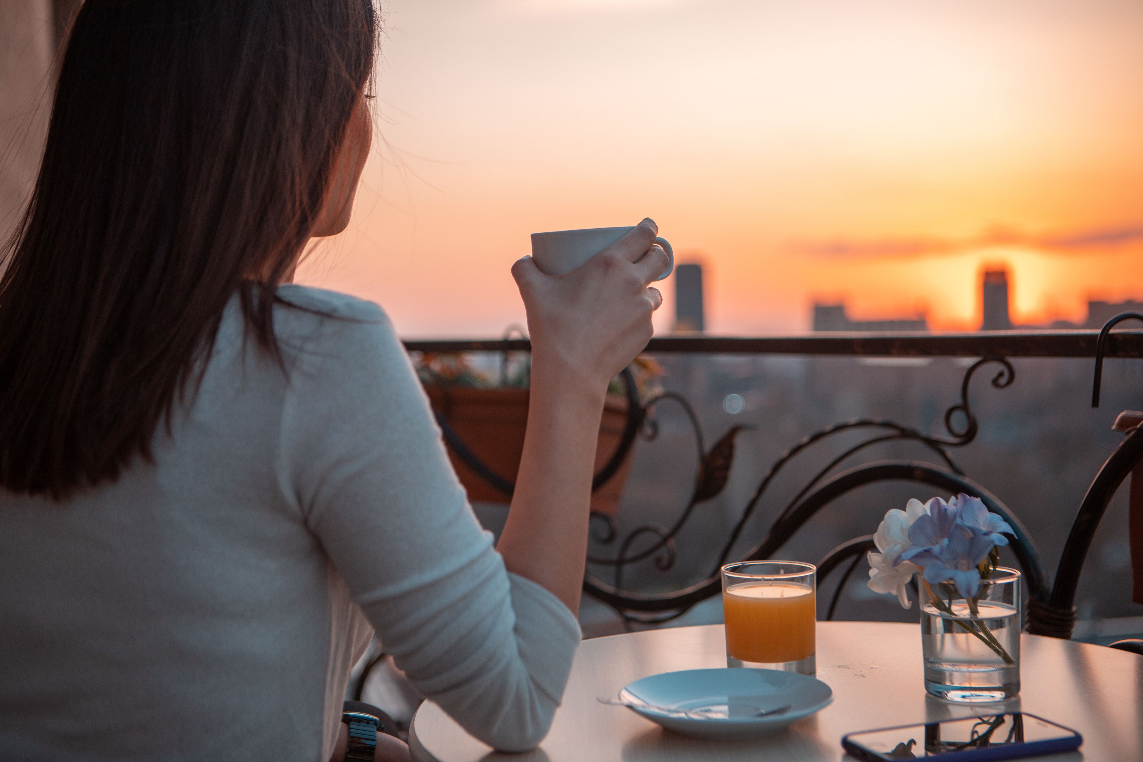 Une personne est assise à une table, devant une tasse et un verre de jus d’orange, et contemple un coucher de soleil avec la silhouette d’une ville en arrière-plan.