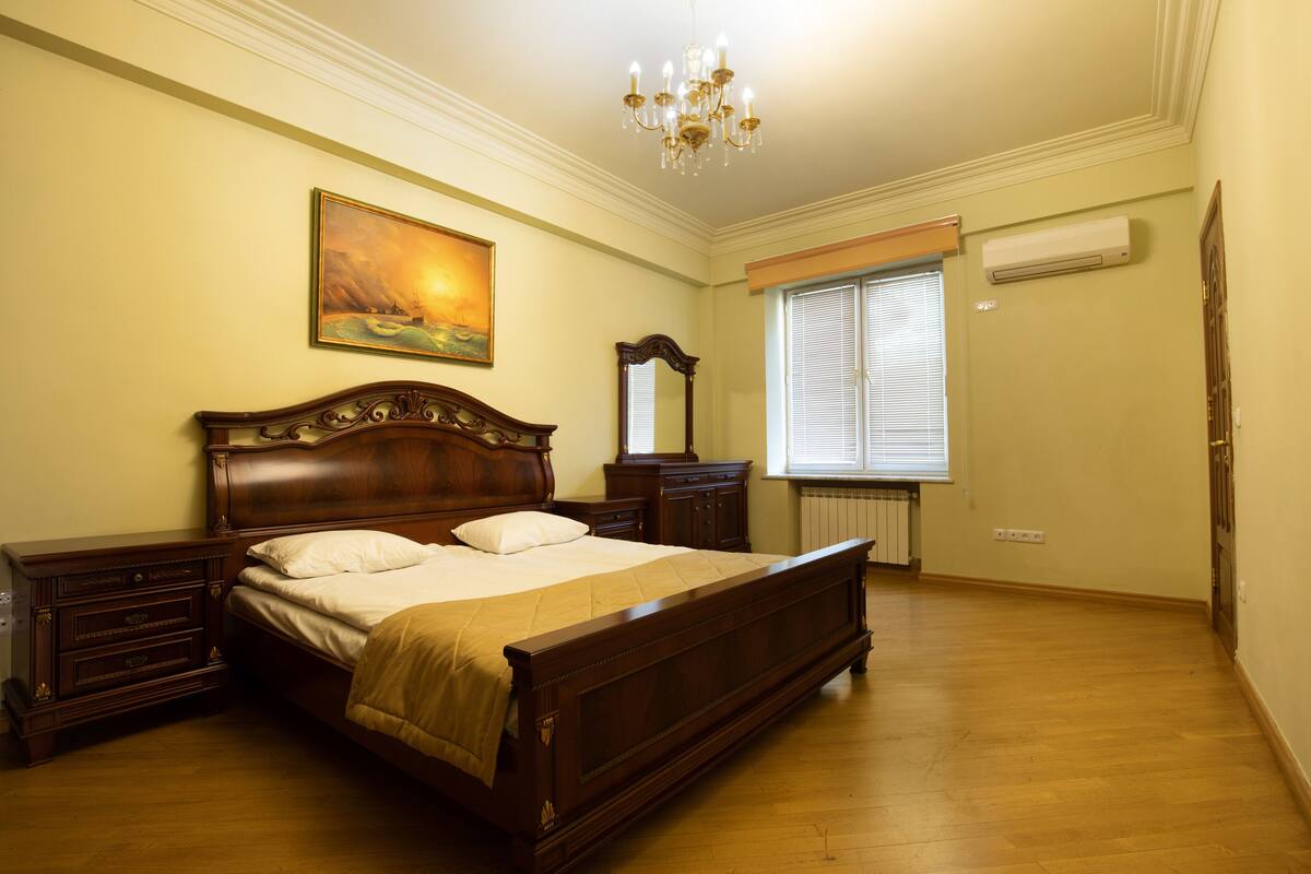 A bedroom with a wooden bed, a nightstand, a dresser, a mirror, a painting on the wall, and a window with blinds.