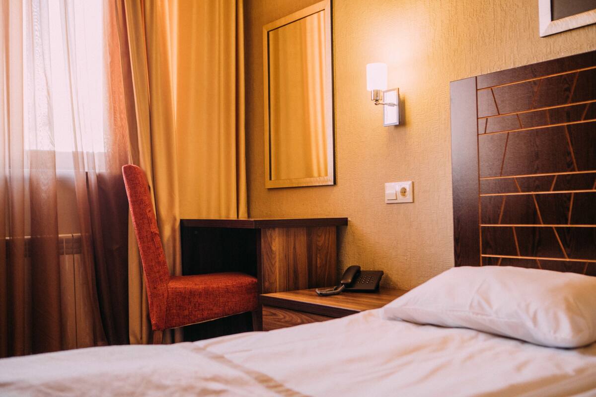 A hotel room with a bed, a chair, a bedside table with a phone, and a wall-mounted light.