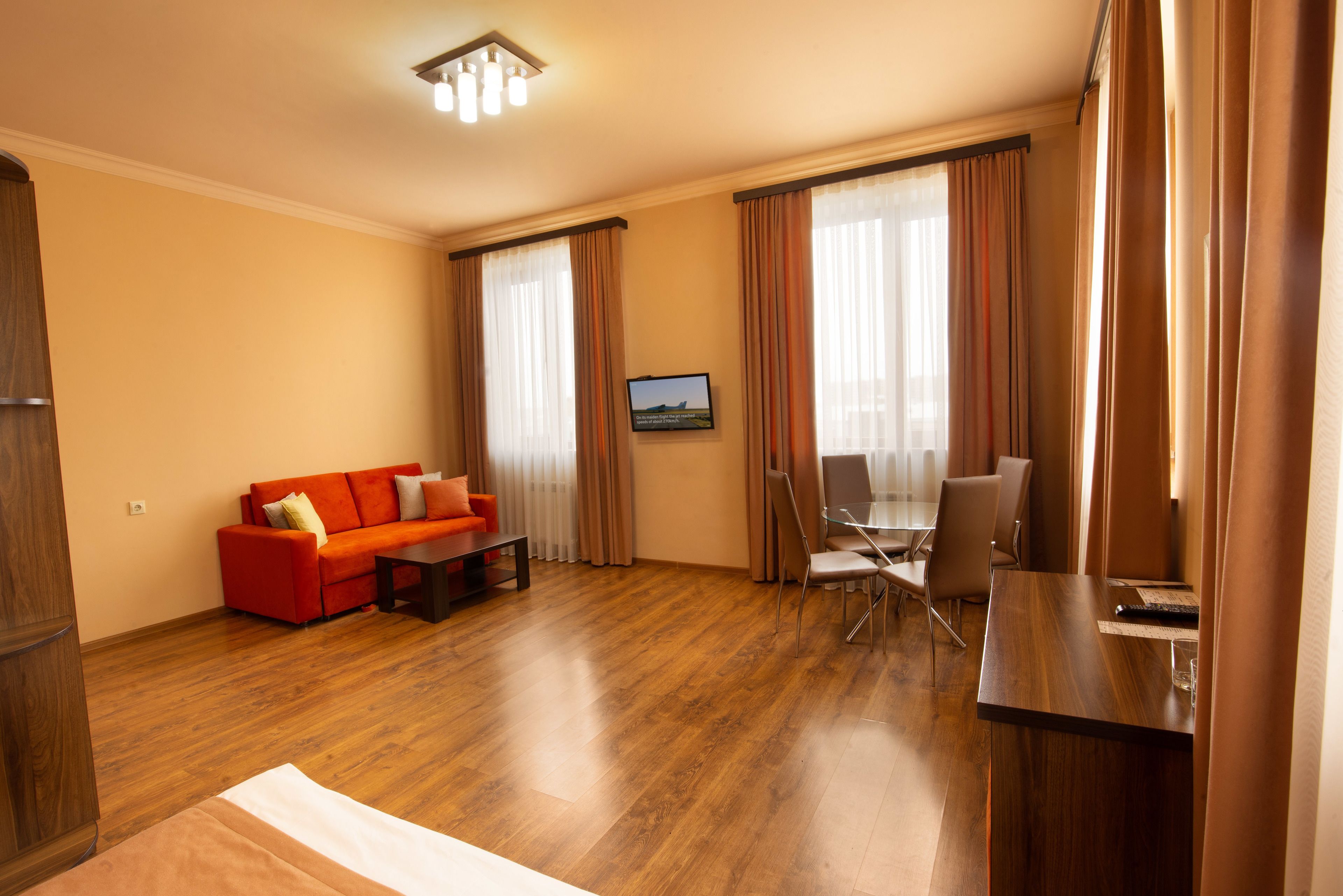 A hotel room with a wooden floor, a red sofa, a dining table with chairs, and a TV on the wall.