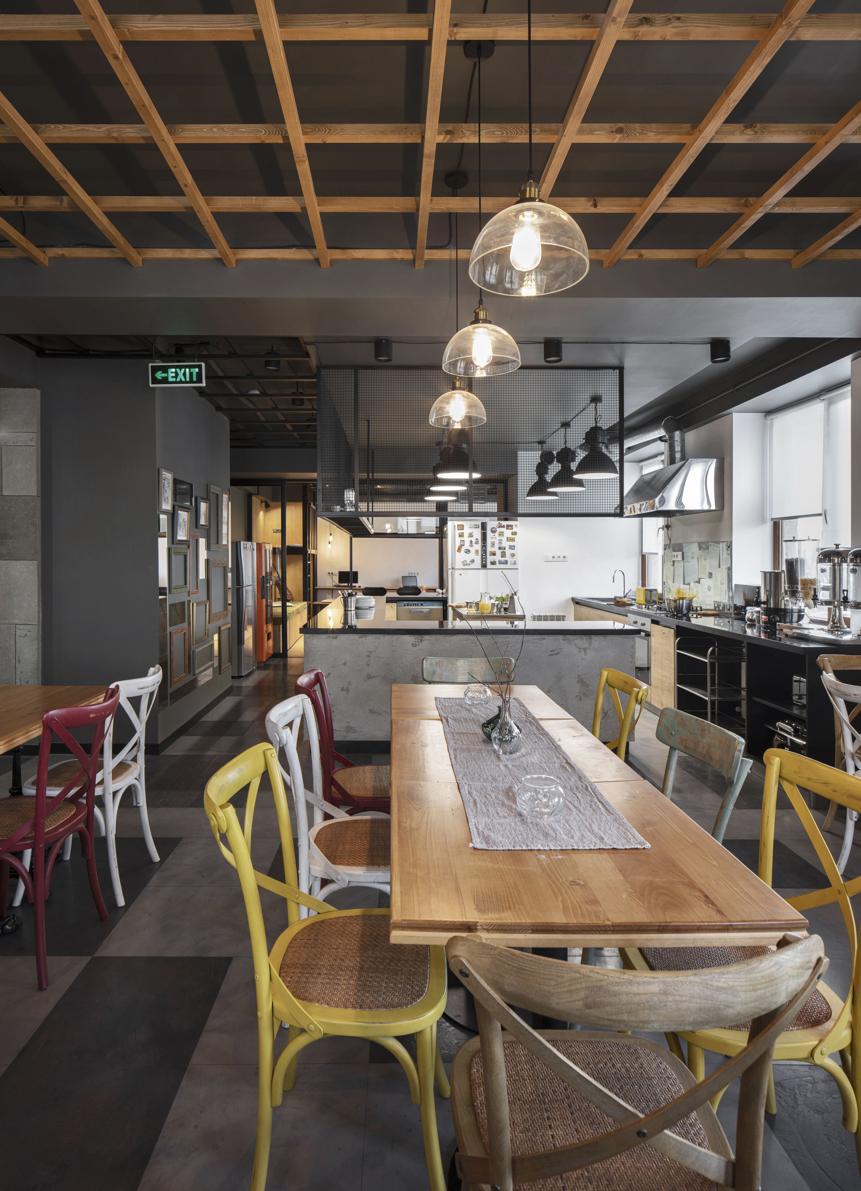 A modern restaurant with a wooden ceiling, a central dining table with chairs, and a kitchen area in the background.