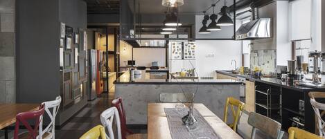 A modern restaurant with a wooden ceiling, a central dining table with chairs, and a kitchen area in the background.