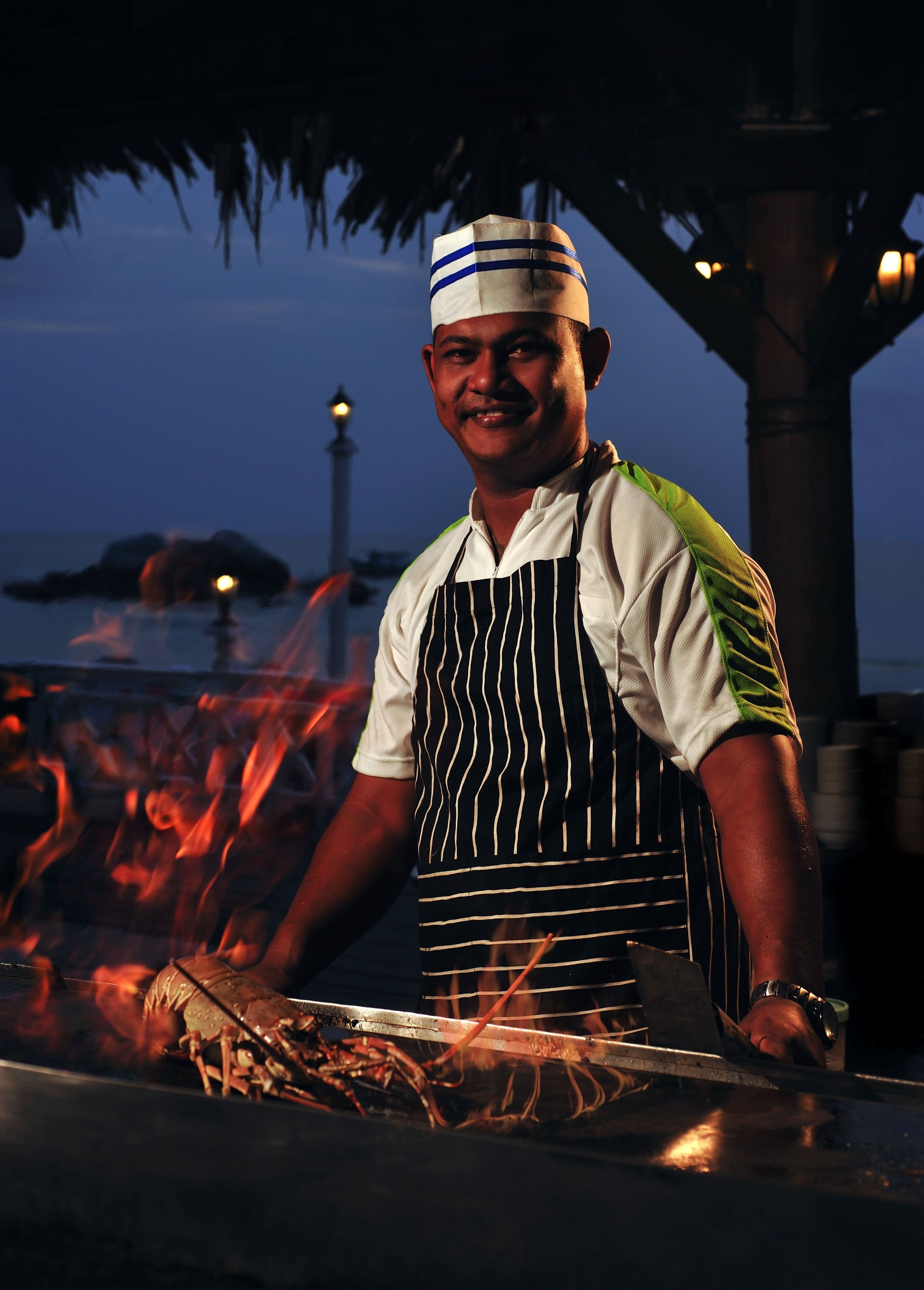 A chef preparing food outdoors at night with a view of the sea and a lighthouse in the background.