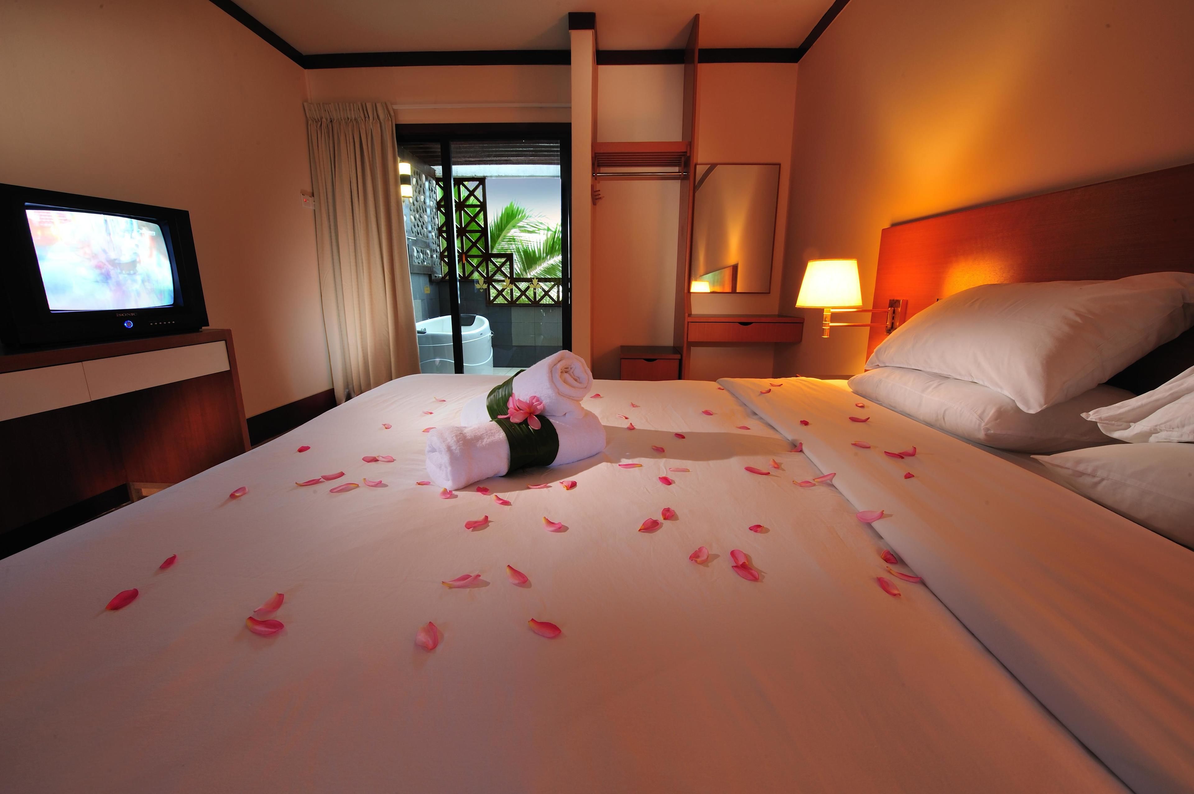 Habitación de hotel con cama, televisión, mesita de noche y balcón con vistas a palmeras.