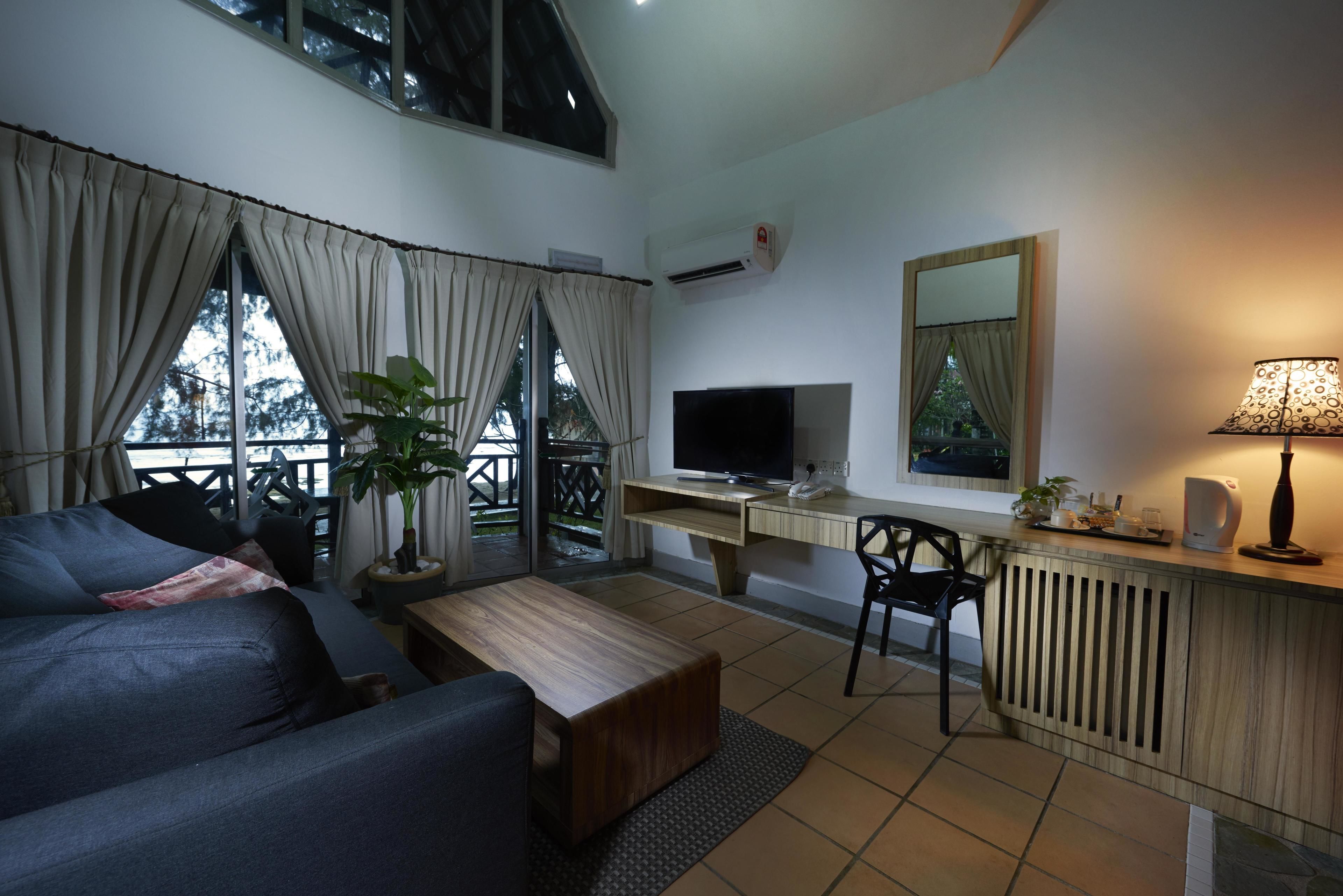 Una sala de estar moderna con un sofá, una mesa de centro, televisión y un escritorio con una computadora. Se ve una ventana con cortinas, una planta en maceta y una lámpara sobre el escritorio.
