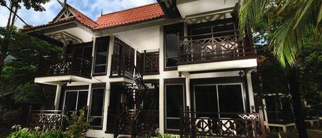 A two-story house with a red-tiled roof, large windows, and a balcony with a wooden railing.
