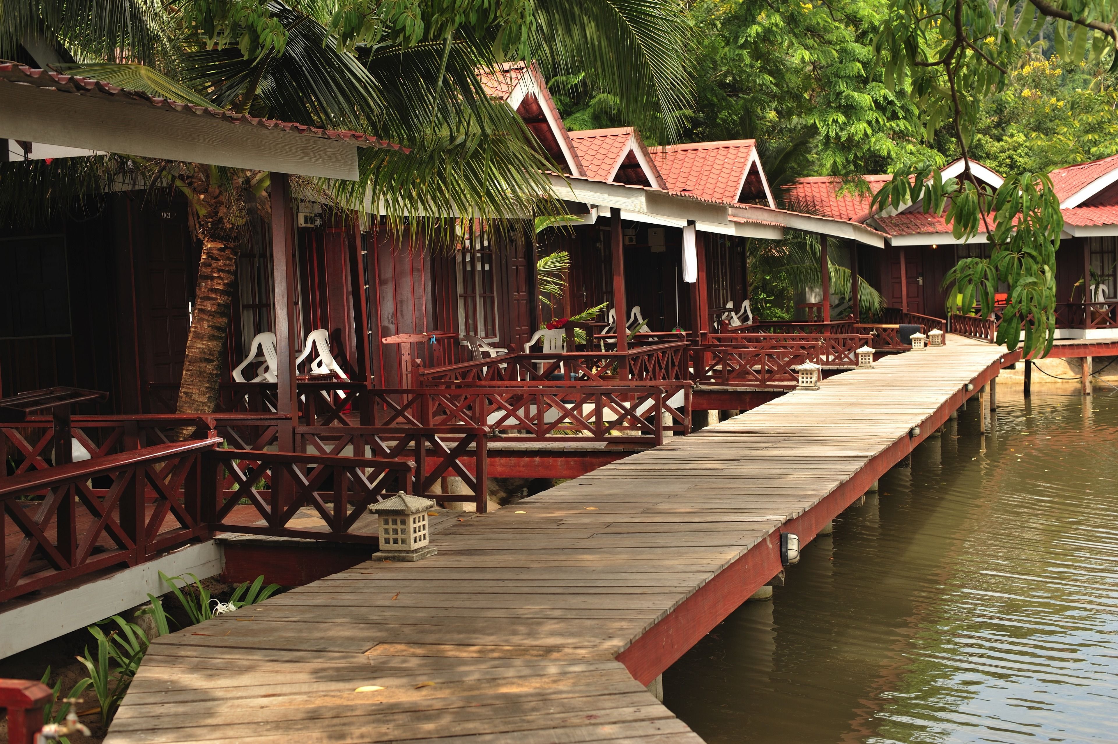 A wooden dock leading to a row of traditional-style buildings with red roofs, surrounded by lush greenery.