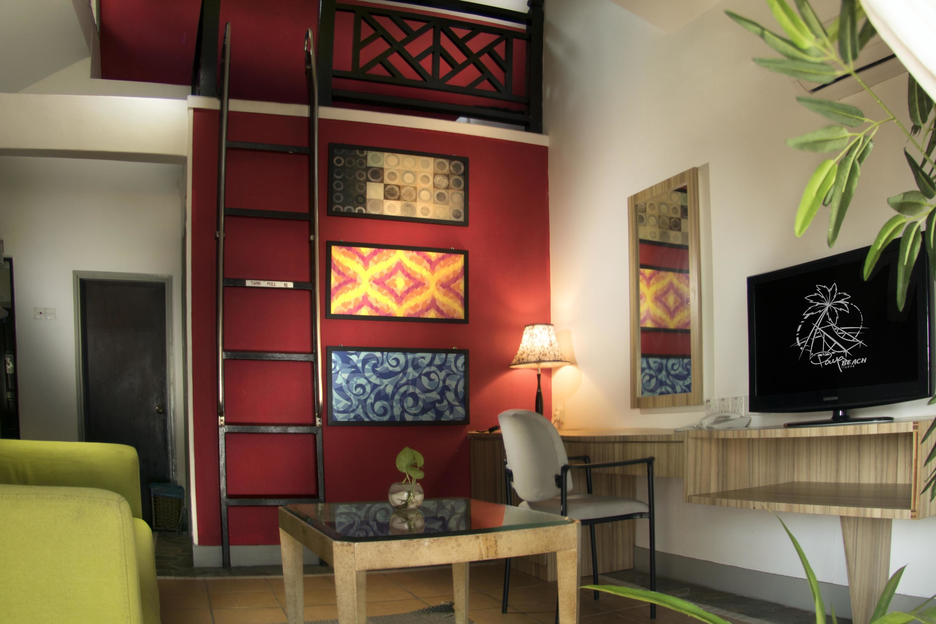 A room with a red accent wall, a TV, a desk, a chair, and a plant.