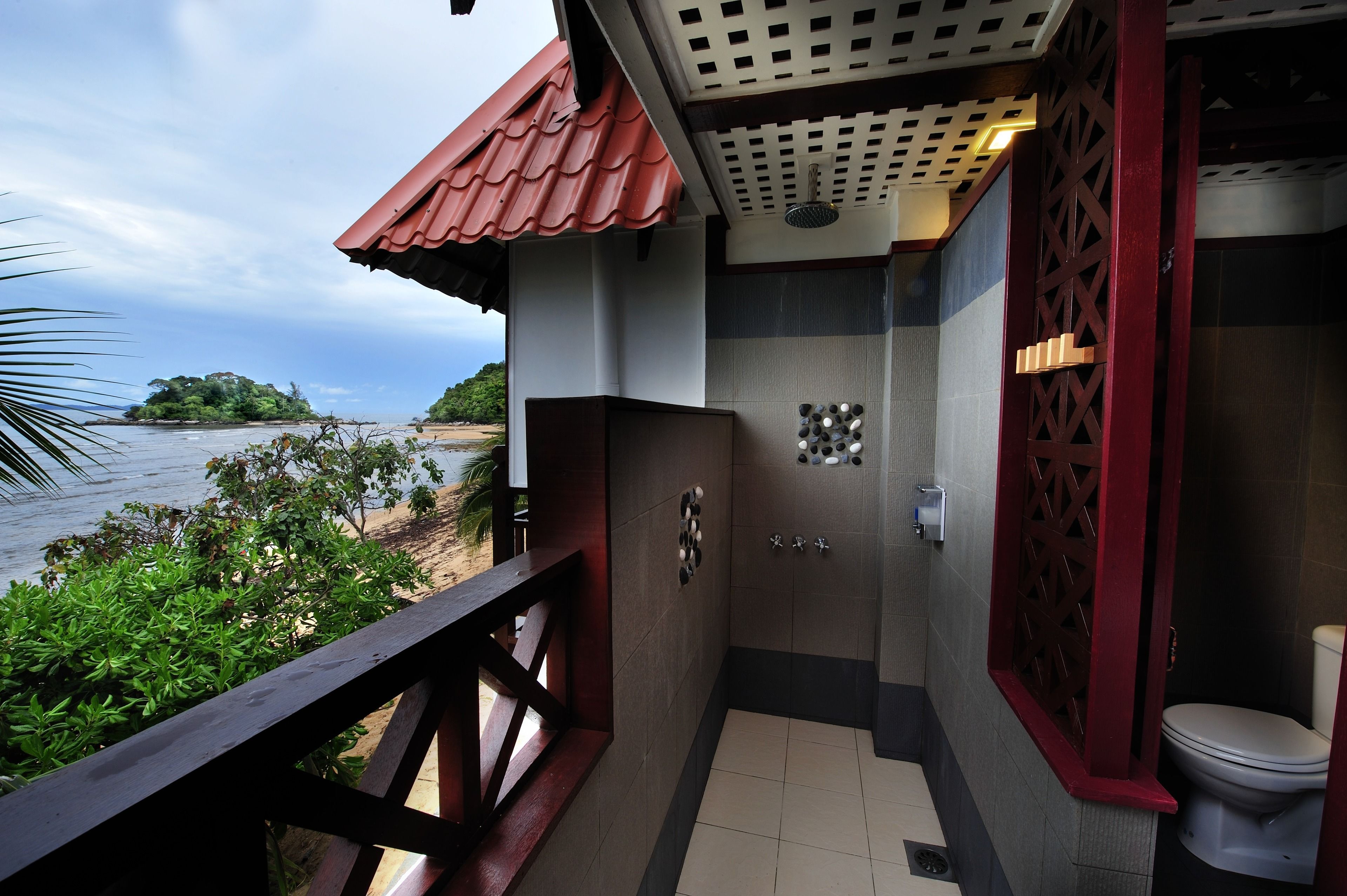 A bathroom with a toilet, a shower, and a sink. There is a balcony with a railing overlooking a beach and ocean.