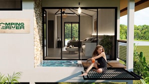 Ett modernt utomhusområde med sittplatser, en glasdörr som leder in till ett vardagsrum, en skylt med texten ”GLAMPING VID FLODEN” och en pool.