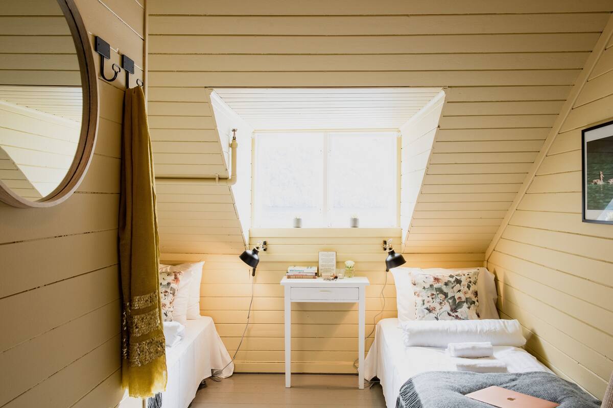 Ein kleiner, gemütlicher Raum mit zwei Betten, einem Tischchen, einem Spiegel und einem Fenster mit Jalousien.