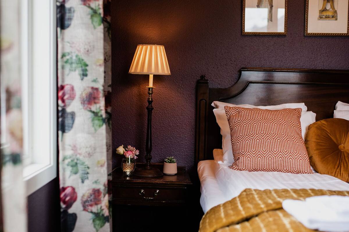 Ein ordentlich bezogenes Bett mit einem gemusterten Überwurf, einer Nachttischlampe und zwei gerahmten Bildern an der Wand.