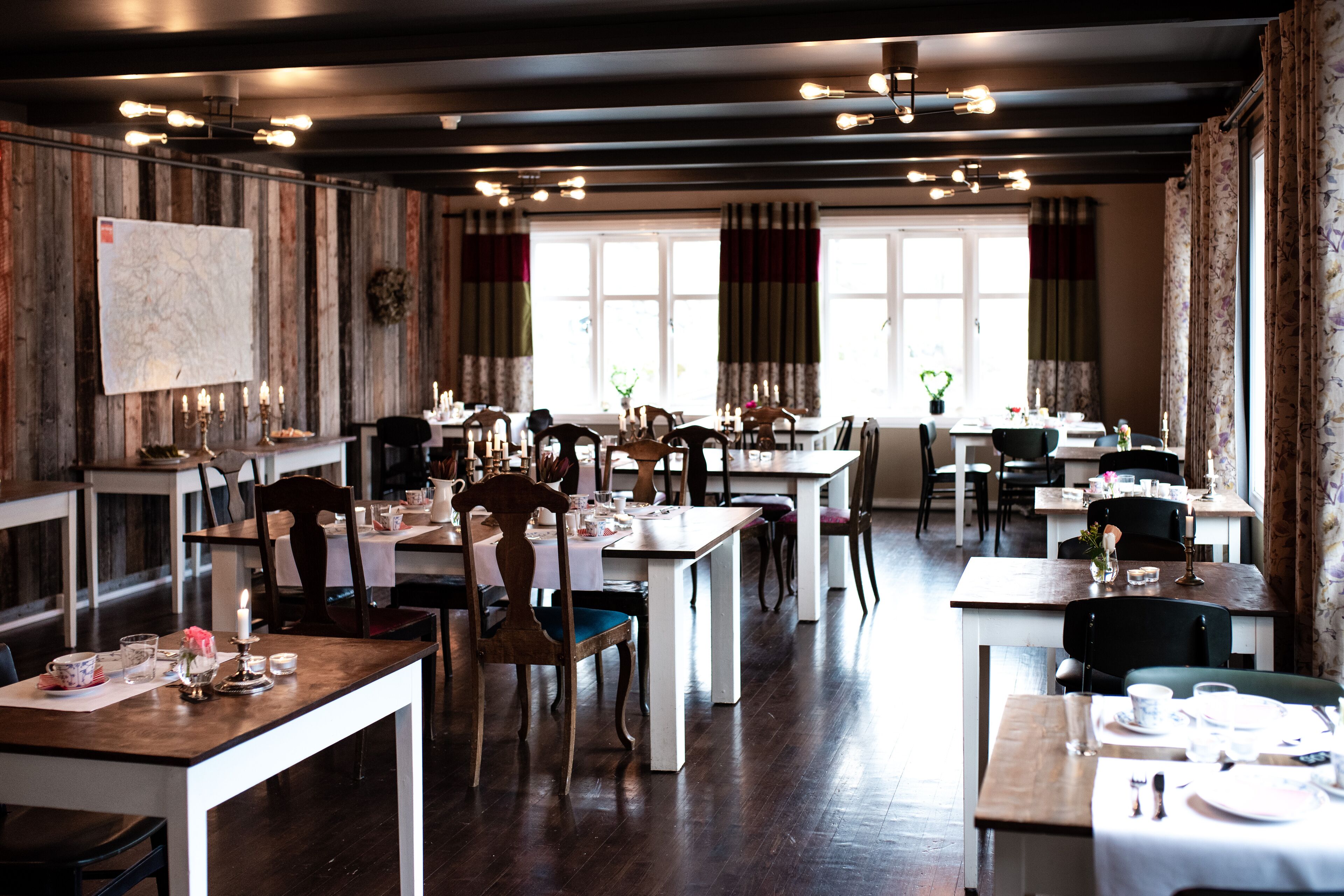 A restaurant with multiple dining tables and chairs, wooden walls, and large windows with curtains.