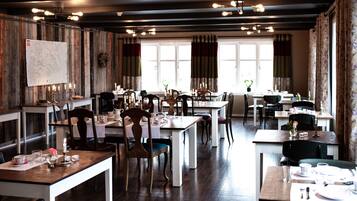 A restaurant with multiple dining tables and chairs, wooden walls, and large windows with curtains.