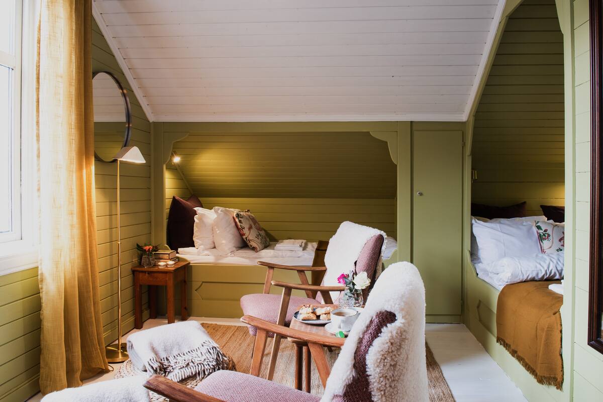 Ein gemütliches Zimmer mit einem Bett, einem Sessel, einem kleinen Tisch mit Tasse und Gebäck sowie einer Lampe.