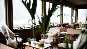 A cozy seating area with a view of the sea, featuring a mix of chairs and sofas, a small table with a bowl of fruit, and various plants. - Fjærland Fjordstove Hotell (Sogndal)