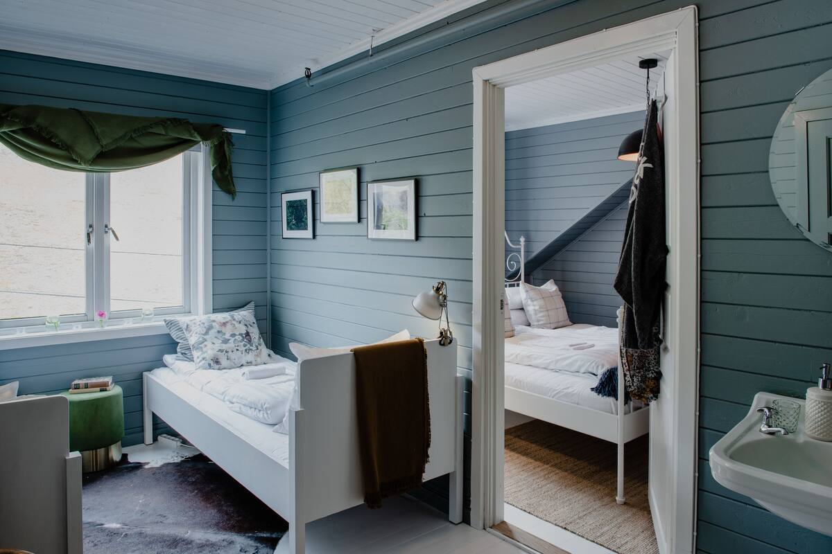 Ein Schlafzimmer mit einem Einzelbett, einem Fenster mit Vorhang, einem Nachttisch mit Lampe und einem Spiegel an der Wand.