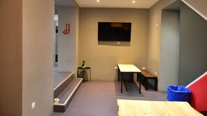 A small, modern room with a TV mounted on the wall, a small table, a bench, and a trash bin. - Zorbas Hotel - Hostel (Athens)