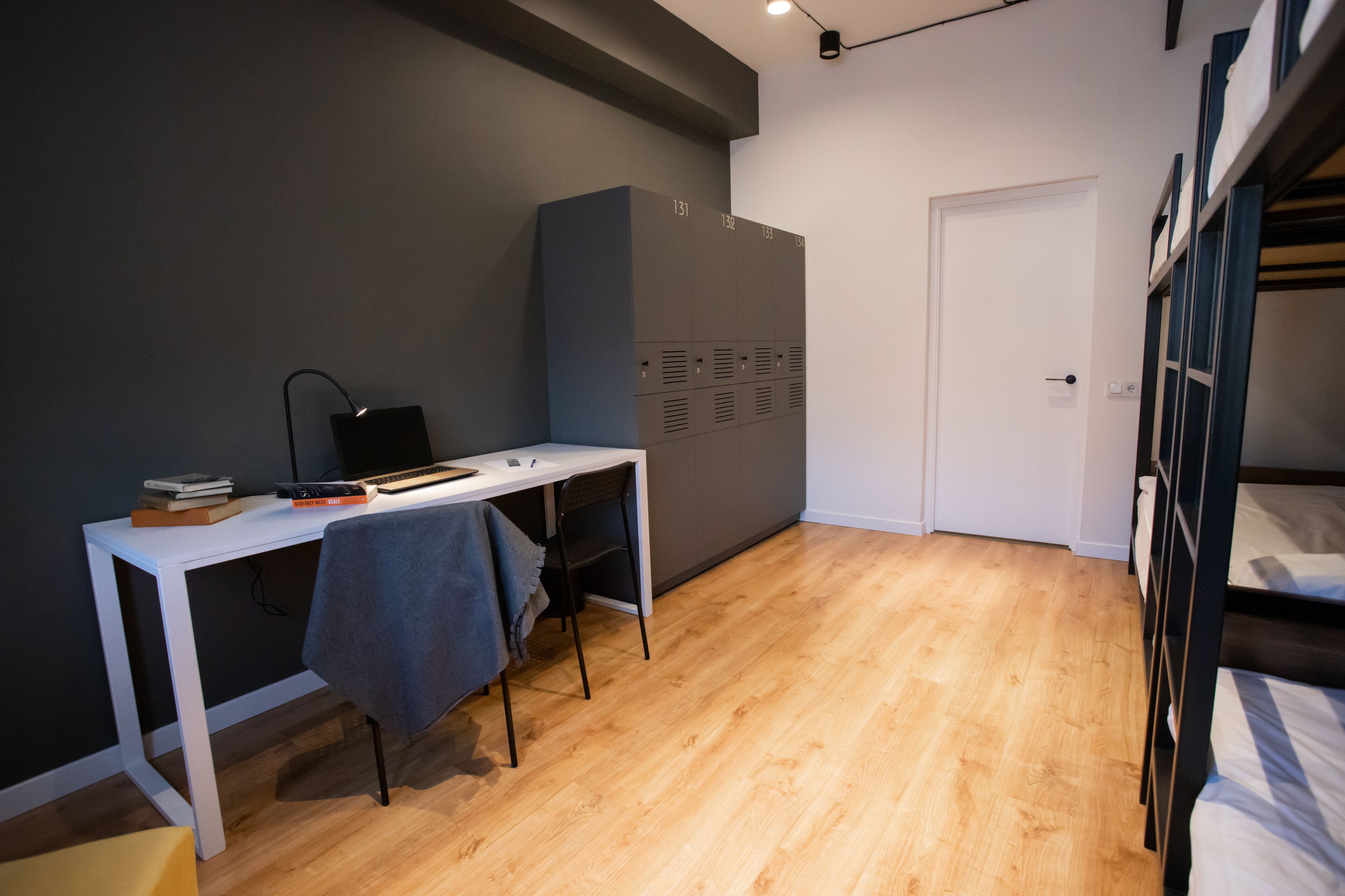 A dormitory room with a bunk bed, a desk with a laptop, a chair, and a wardrobe with lockers.