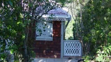 A small wooden cabin with a white door and a decorative window grille, surrounded by dense foliage.