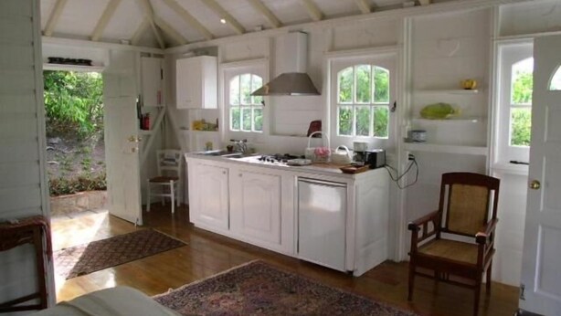 A compact kitchen with white cabinetry, a central island, and a dining area with a chair. - Fire Fly Cottage (Westbury)