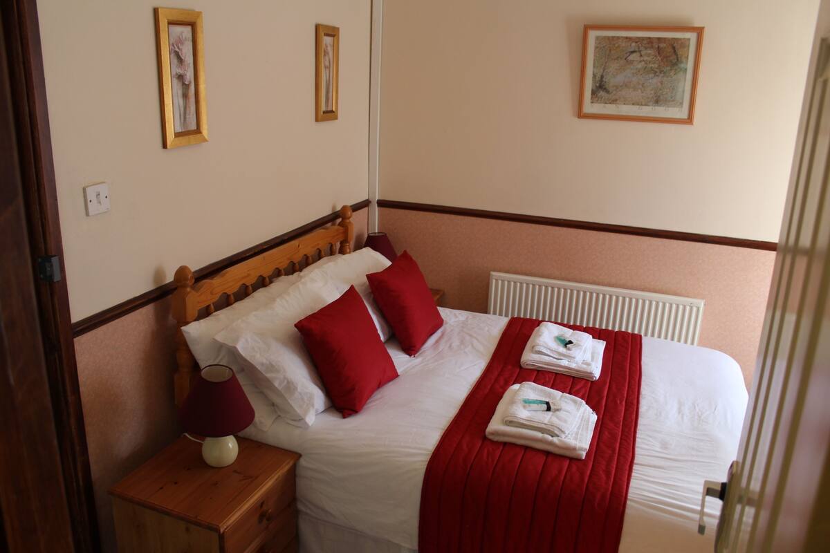 A neatly made bed with red pillows, a red blanket, and a folded towel on a wooden nightstand.