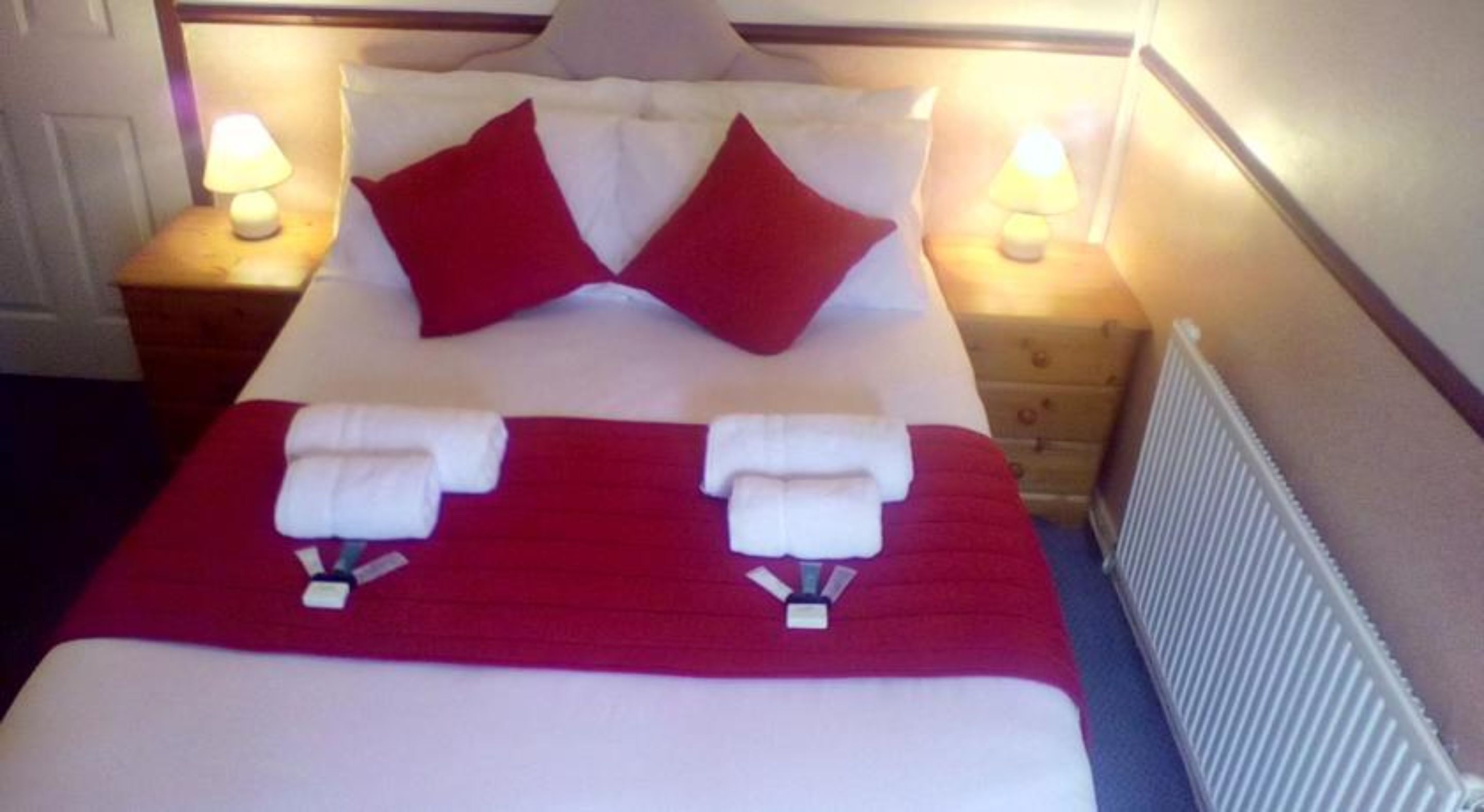 A neatly made bed with a red blanket and pillows, two towels on the bed, and bedside lamps on wooden nightstands.