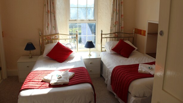 A bedroom with two beds, each with a red blanket and pillow, a window with floral curtains, and a nightstand with a lamp. - The Middlegate Hotel (Pembroke)