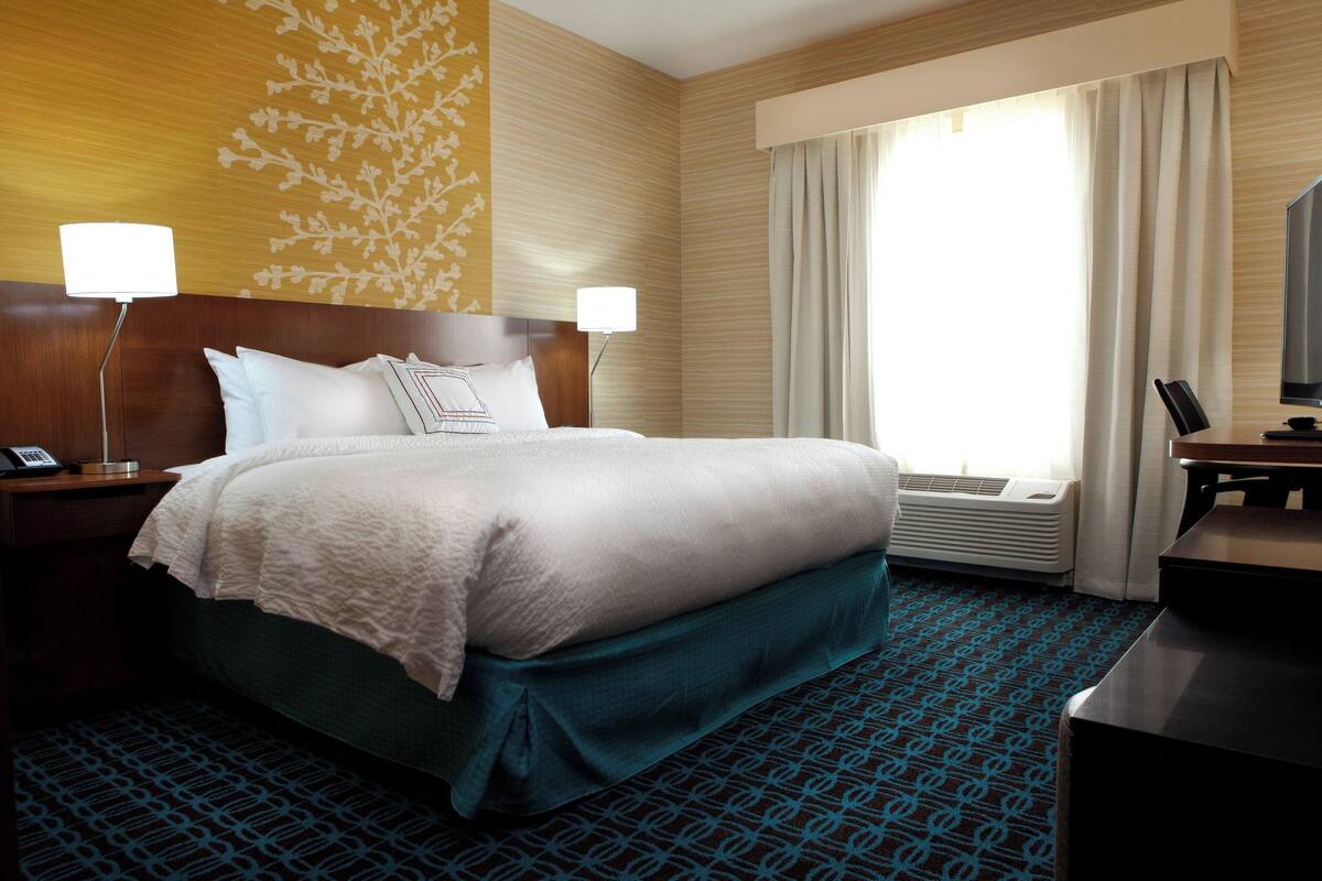 Ein Hotelzimmer mit einem großen Bett, zwei Nachttischen, einem Schreibtisch und einem großen Fenster mit Vorhängen.