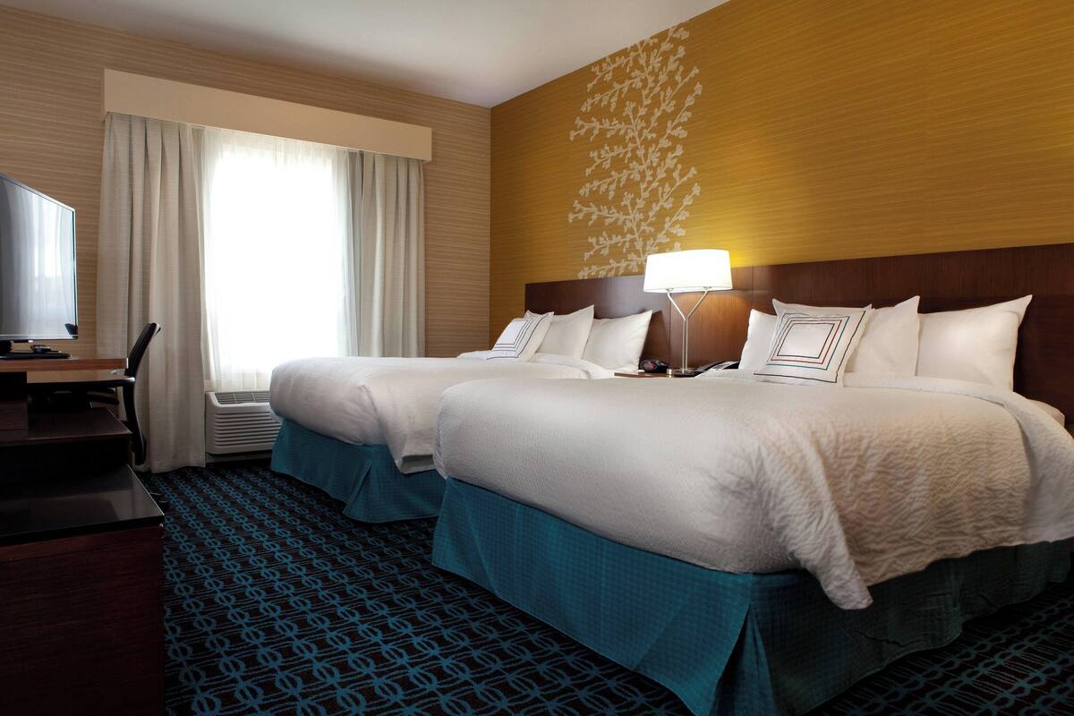 Ein Hotelzimmer mit zwei Betten, einem Schreibtisch, einem Fernseher und einem großen Fenster mit Vorhängen.