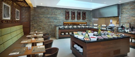 Um buffet de café da manhã moderno com diversas opções de comida, área de estar com mesas e cadeiras, e um chef preparando alimentos ao fundo.