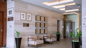 Un vestíbulo moderno con suelo de madera, dos sillones beige, un mostrador de recepción, obras de arte enmarcadas y señales indicativas para salas de reuniones y estacionamiento.