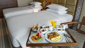 Habitación de hotel con una cama bien hecha, una mesita de noche con una lámpara y una bandeja de desayuno con diversos alimentos y bebidas.