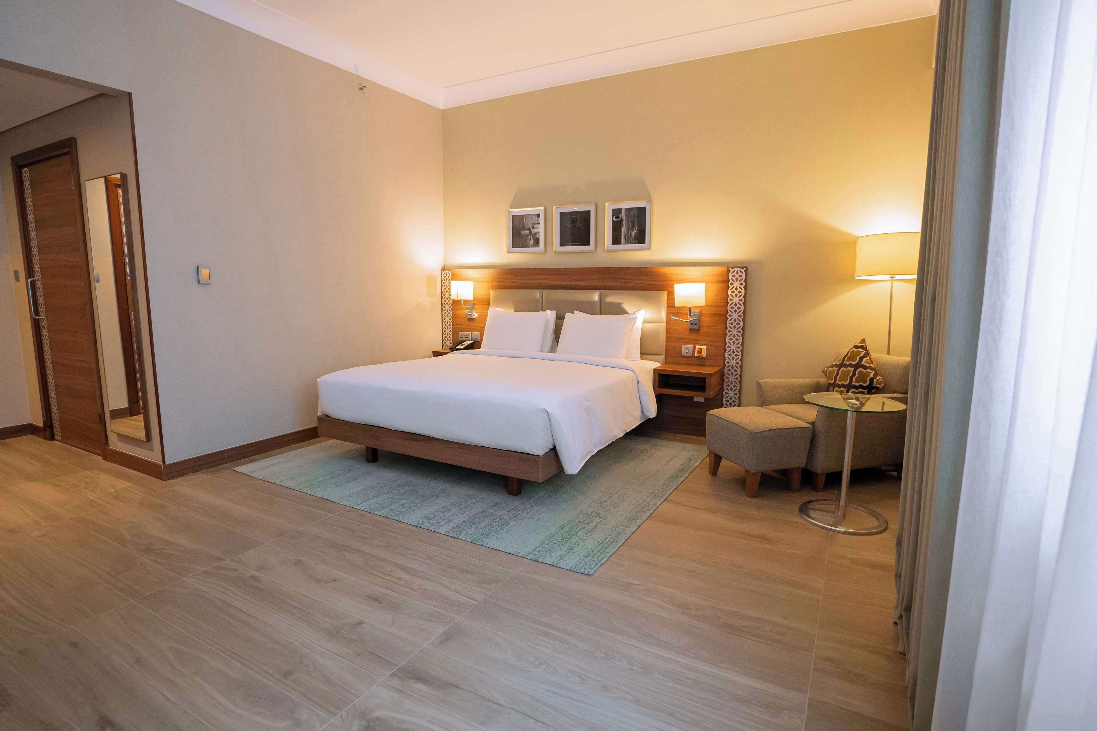 Una habitación de hotel con cama, mesitas de noche, una silla y una pequeña mesa con lámpara.