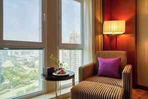 Une chambre d’hôtel avec une grande fenêtre, un fauteuil à rayures agrémenté d’un coussin violet, une petite table avec une plante et un vase, et un lampadaire.