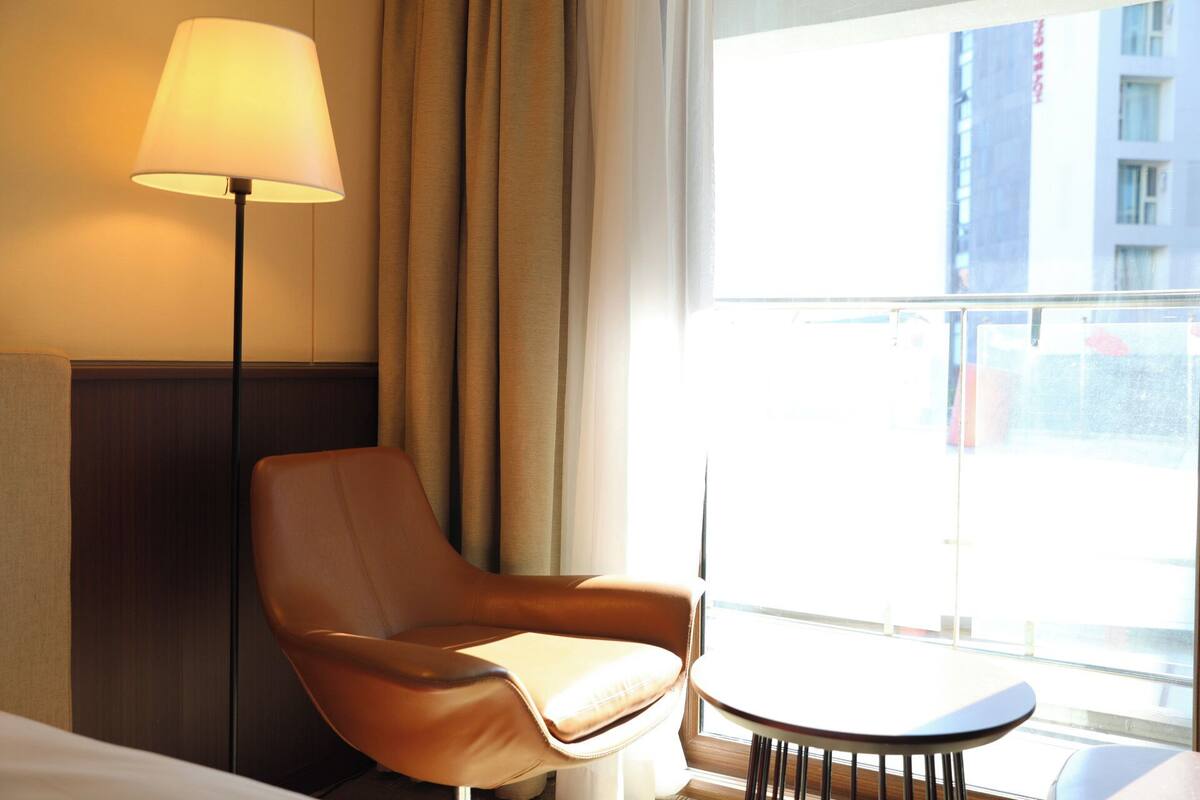 Une chambre d’hôtel avec un fauteuil en cuir, une petite table ronde, un lampadaire et une grande fenêtre donnant sur des immeubles.