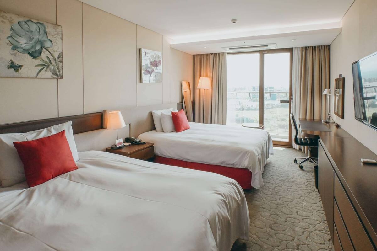 Une chambre d’hôtel avec deux lits, un bureau, une chaise, une télévision et une grande fenêtre donnant sur la ville.