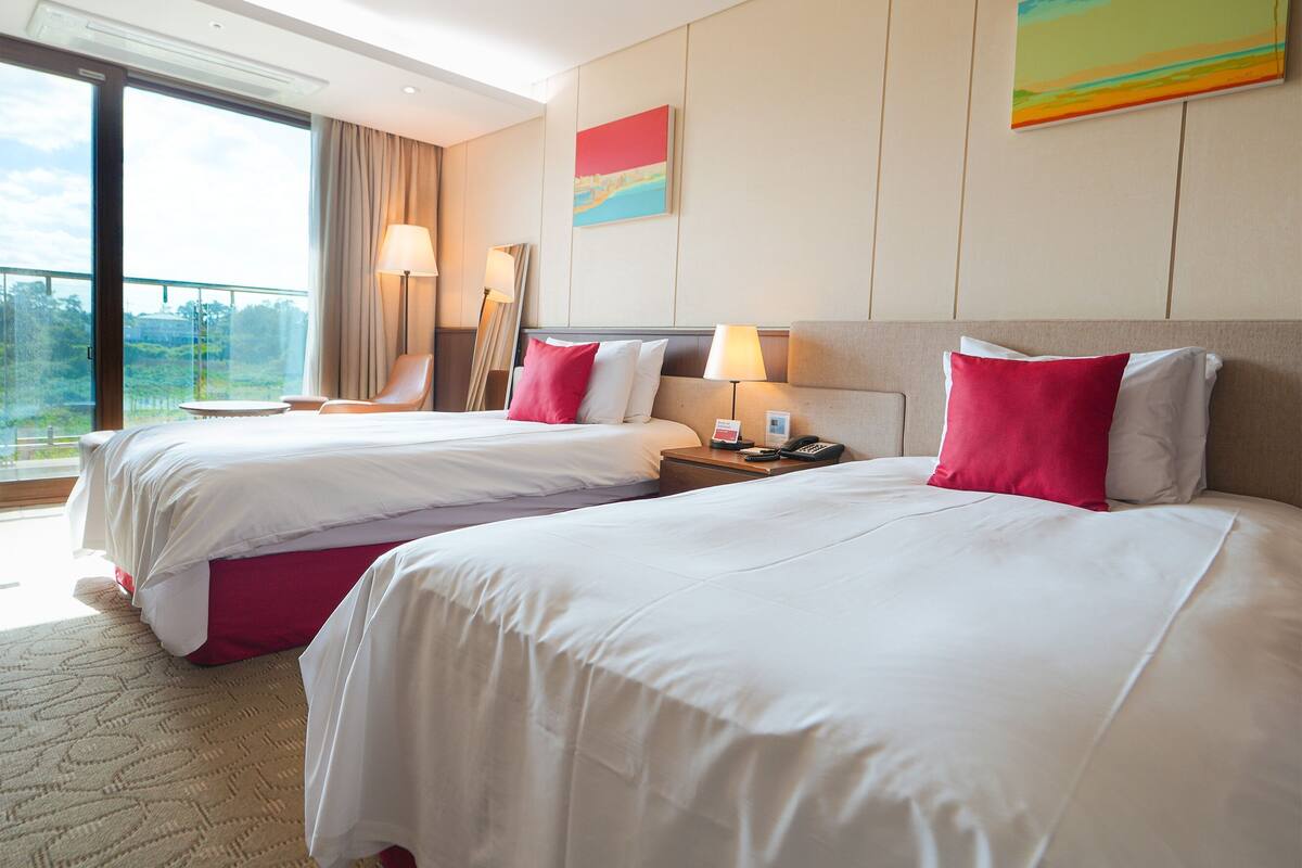 Une chambre d’hôtel avec deux lits, une table de chevet, une lampe, un tableau et une vue sur l’extérieur.