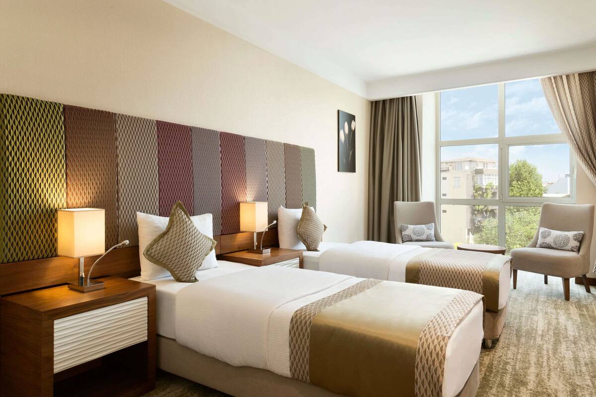 Quarto de hotel com duas camas, uma janela grande com cortinas, cabeceira com padrão decorativo e vista para prédios.