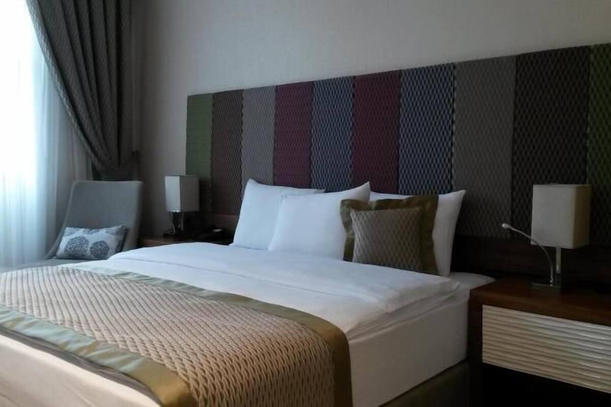 Quarto de hotel com uma cama grande, duas mesinhas de cabeceira e uma janela com cortinas.