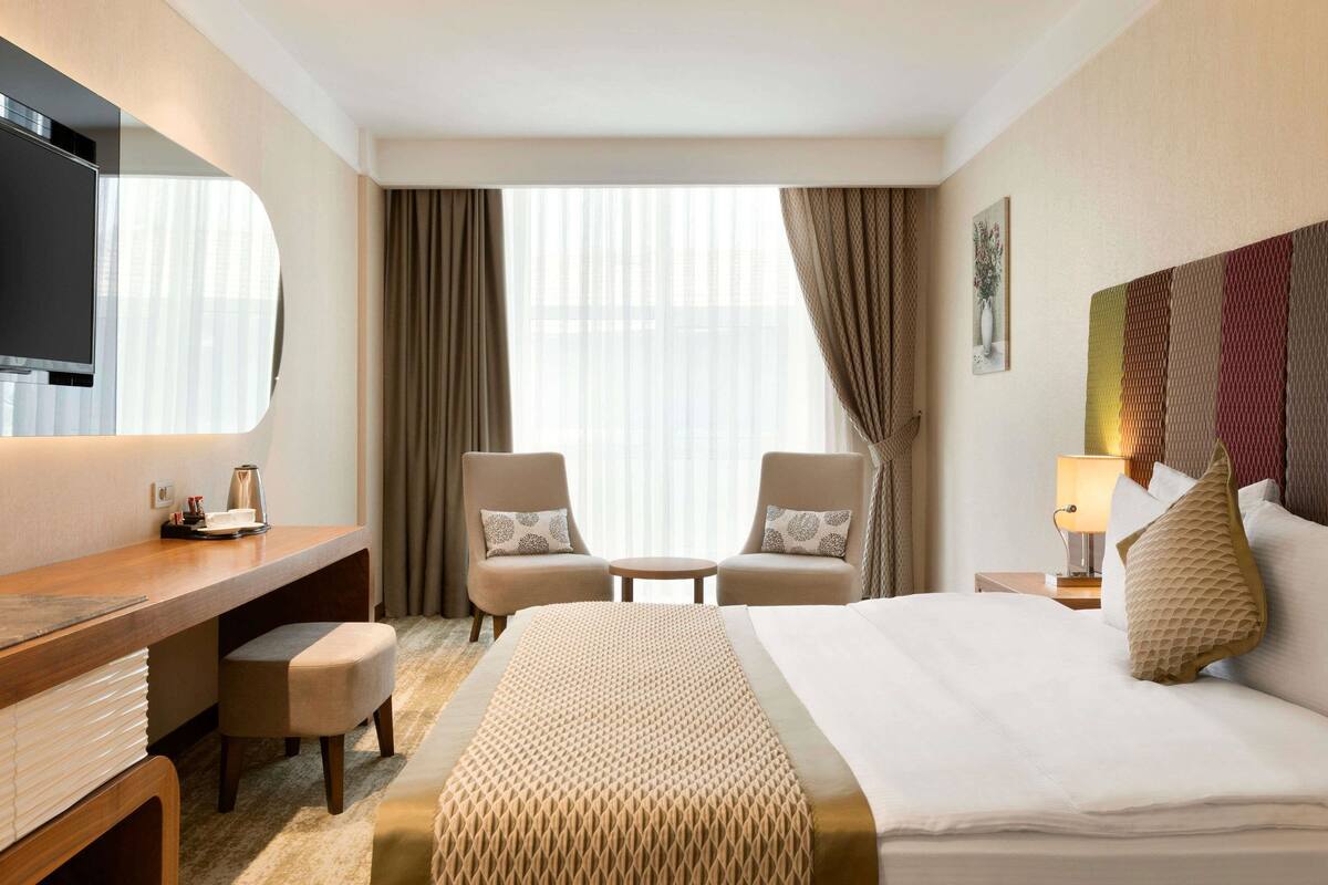Quarto de hotel com cama, escrivaninha, duas cadeiras, televisão e janela com cortinas.