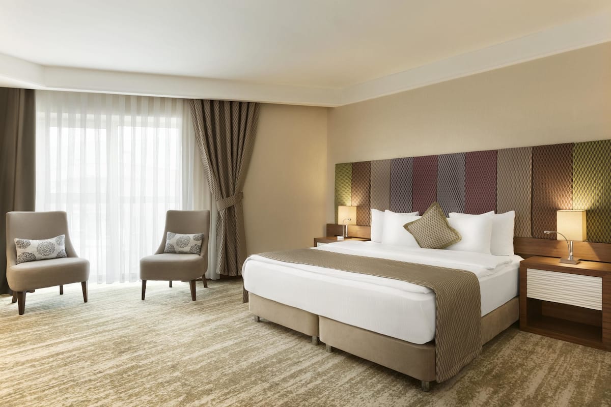 Quarto de hotel com uma cama grande, duas poltronas, uma mesa de cabeceira com abajur e vista para o exterior através de cortinas finas.