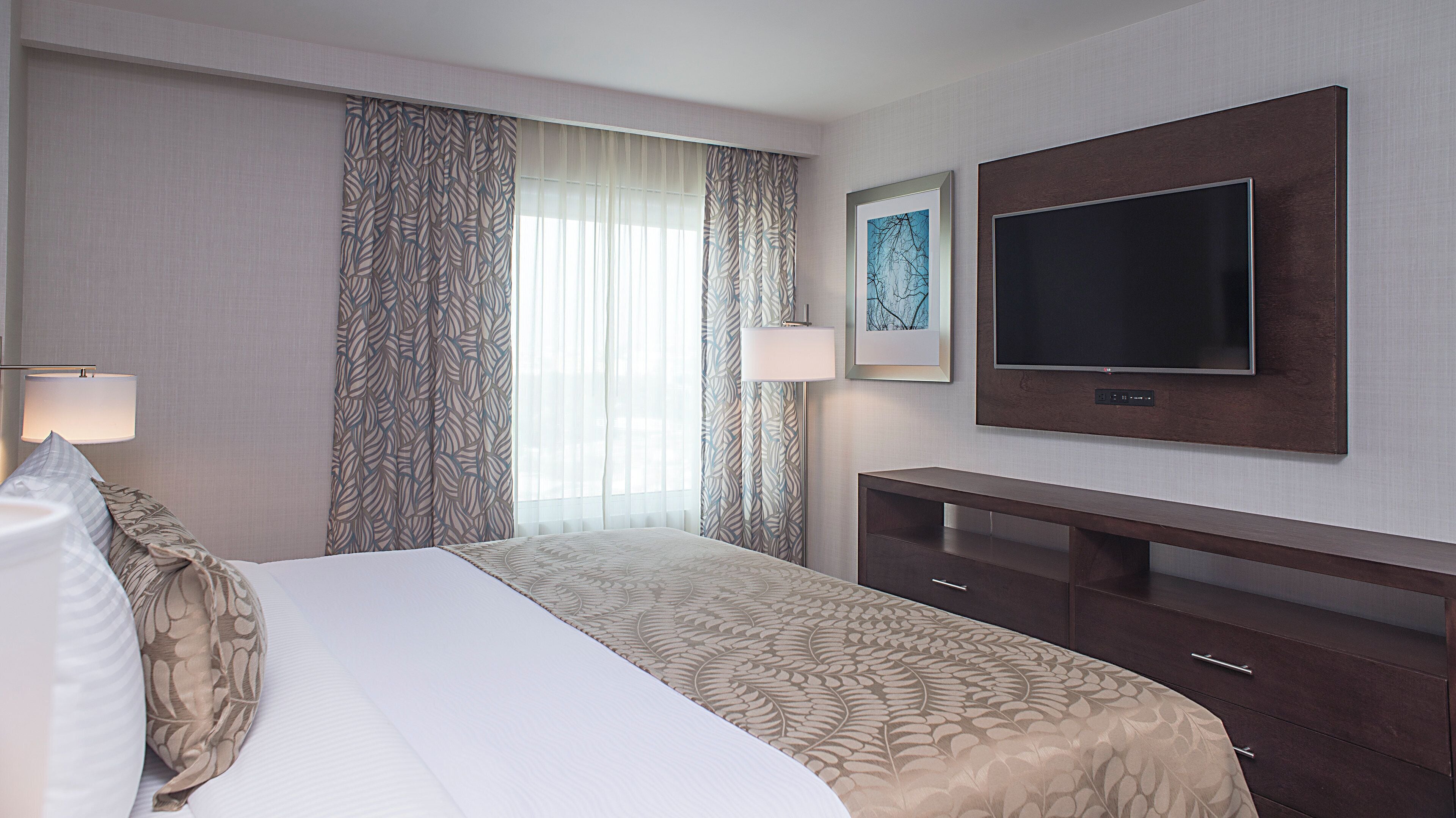 Een hotelkamer met een bed, een flatscreen televisie aan de muur, een nachtkastje en een groot raam met dunne gordijnen.